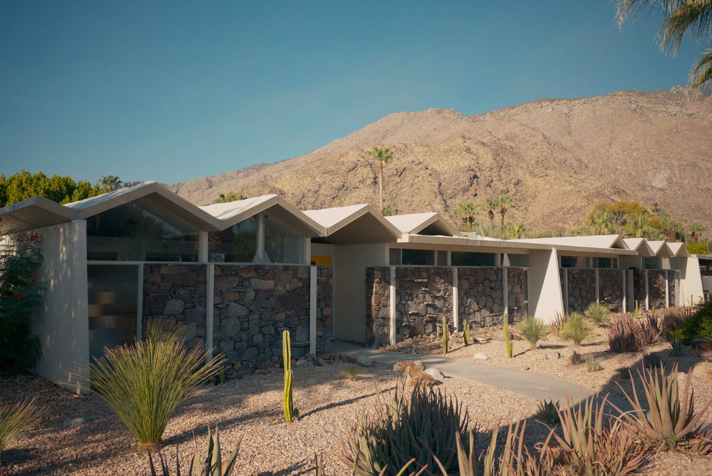 A mid-century modern house with a stone wall and folded roofline below desert mountains